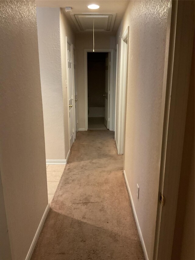 hall featuring baseboards, a textured wall, attic access, and light colored carpet