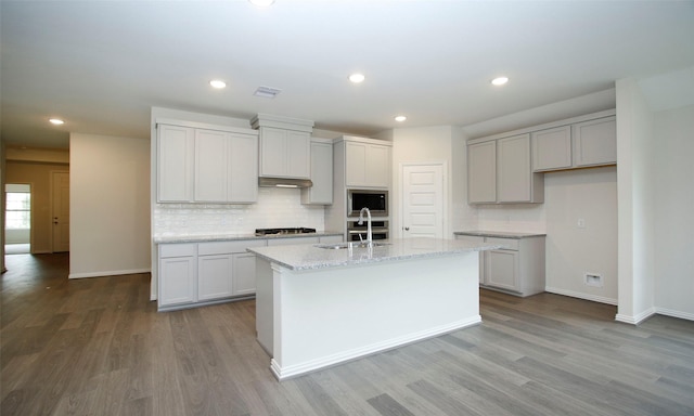 kitchen with sink, light hardwood / wood-style floors, an island with sink, light stone countertops, and built in microwave