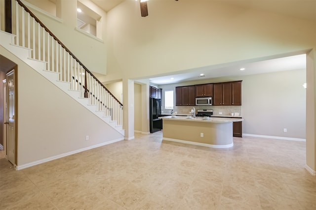 kitchen with dark brown cabinets, appliances with stainless steel finishes, an island with sink, a towering ceiling, and ceiling fan