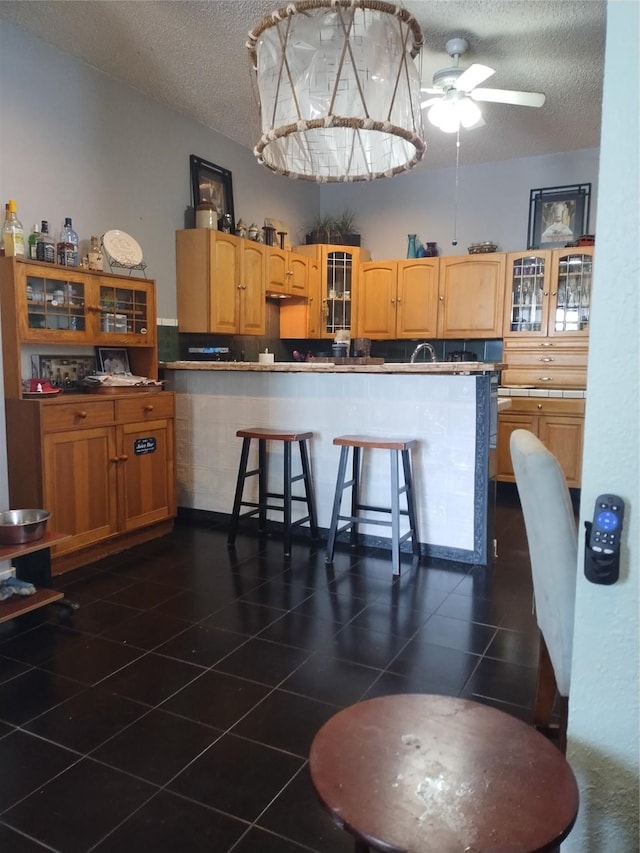kitchen featuring ceiling fan, a kitchen breakfast bar, a textured ceiling, and glass insert cabinets
