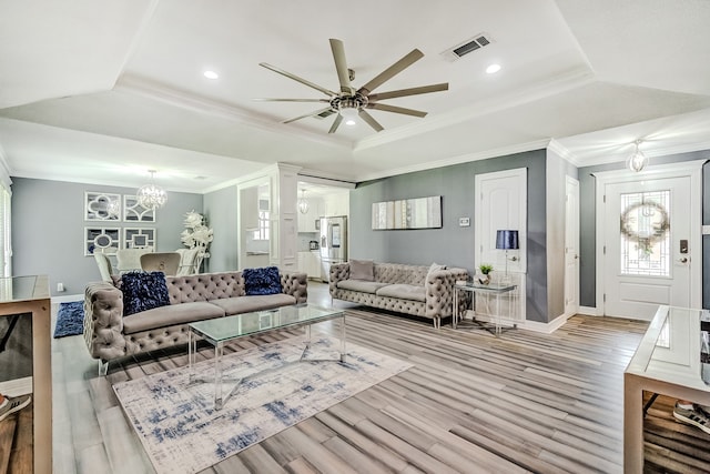 living room with light wood-type flooring, a tray ceiling, ceiling fan with notable chandelier, and ornamental molding