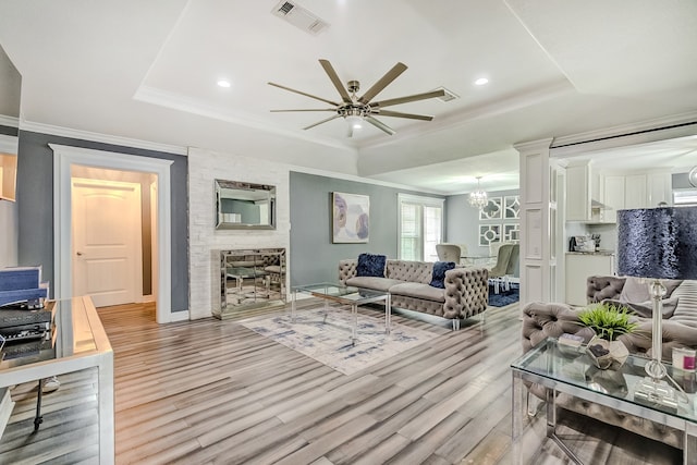 living room with ceiling fan with notable chandelier, a raised ceiling, ornamental molding, and light hardwood / wood-style flooring