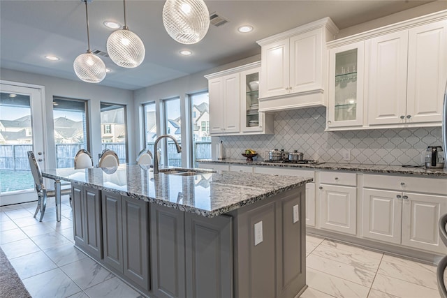 kitchen with light stone countertops, a kitchen island with sink, sink, pendant lighting, and white cabinetry