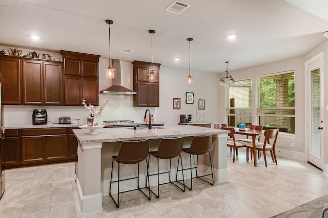 kitchen with light tile patterned flooring, an island with sink, wall chimney exhaust hood, and decorative light fixtures