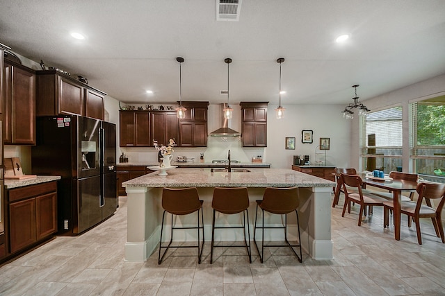 kitchen featuring wall chimney range hood, pendant lighting, an island with sink, black fridge, and dark brown cabinetry