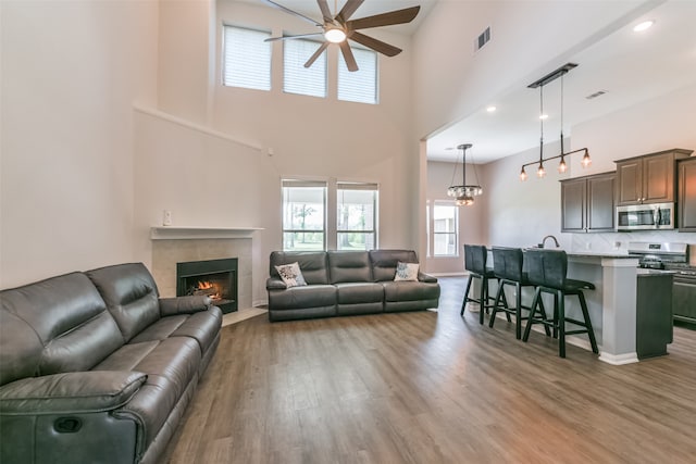 living room featuring a high ceiling, ceiling fan with notable chandelier, light hardwood / wood-style floors, and a tiled fireplace