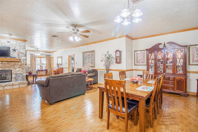 dining area with a stone fireplace, crown molding, a textured ceiling, light wood-type flooring, and ceiling fan with notable chandelier