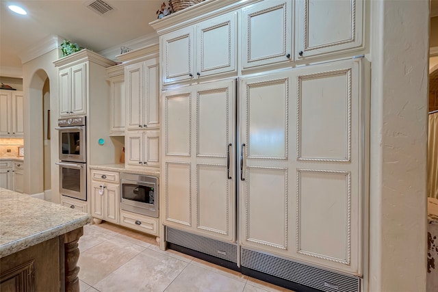 kitchen with appliances with stainless steel finishes, backsplash, light tile patterned floors, cream cabinets, and ornamental molding