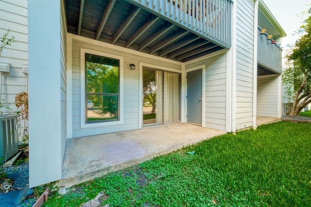 view of exterior entry with a balcony, a yard, a patio area, and central AC