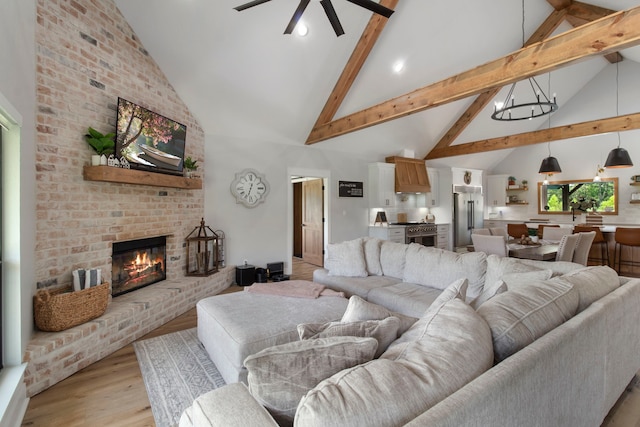 living room with a brick fireplace, ceiling fan with notable chandelier, high vaulted ceiling, beam ceiling, and light hardwood / wood-style flooring