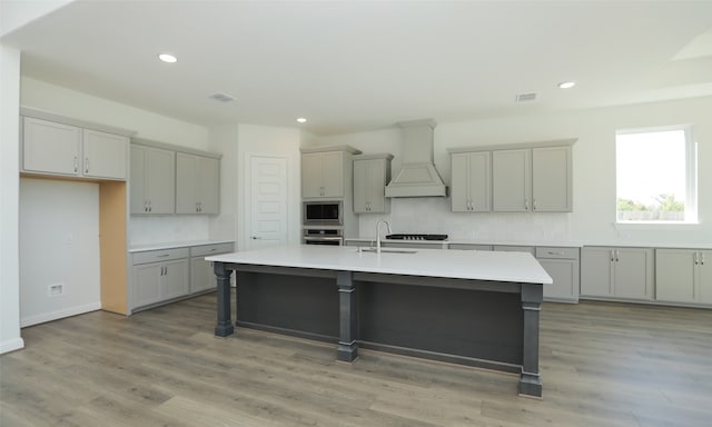 kitchen featuring stainless steel appliances, an island with sink, gray cabinets, custom range hood, and light wood-type flooring