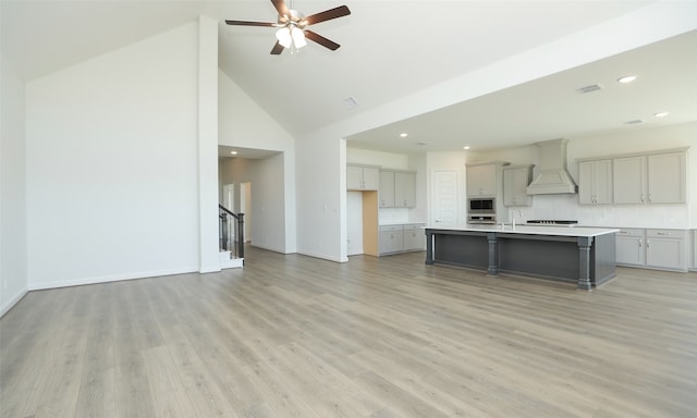 kitchen featuring custom exhaust hood, a kitchen island with sink, light hardwood / wood-style flooring, high vaulted ceiling, and stainless steel microwave