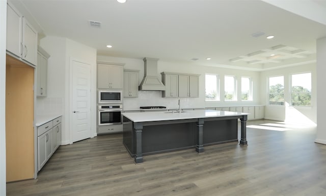 kitchen with dark wood-type flooring, coffered ceiling, premium range hood, a kitchen island with sink, and appliances with stainless steel finishes