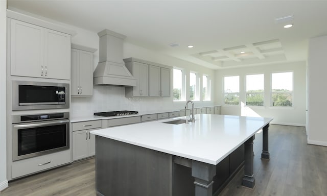 kitchen featuring coffered ceiling, custom exhaust hood, stainless steel appliances, a kitchen island with sink, and light hardwood / wood-style floors