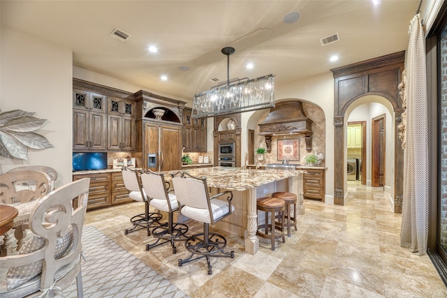 kitchen featuring custom range hood, hanging light fixtures, built in appliances, a large island with sink, and light stone counters