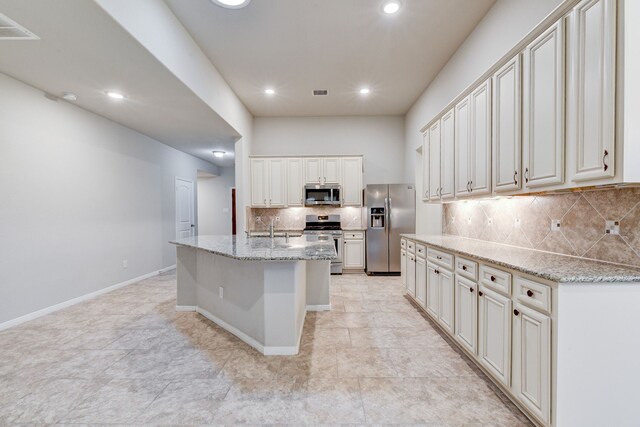 kitchen with light stone counters, stainless steel appliances, an island with sink, and decorative backsplash