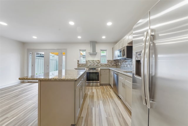 kitchen featuring tasteful backsplash, stainless steel appliances, wall chimney exhaust hood, light stone counters, and a kitchen island