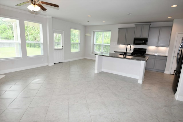 kitchen with gray cabinetry, backsplash, dark stone counters, a kitchen island with sink, and black appliances