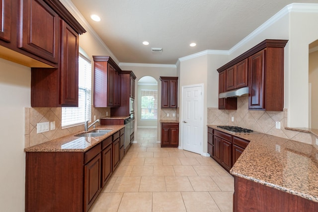 kitchen with arched walkways, under cabinet range hood, stainless steel appliances, a sink, and light stone countertops