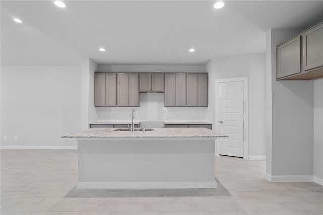 kitchen with light stone countertops, light wood-type flooring, tasteful backsplash, a kitchen island with sink, and sink