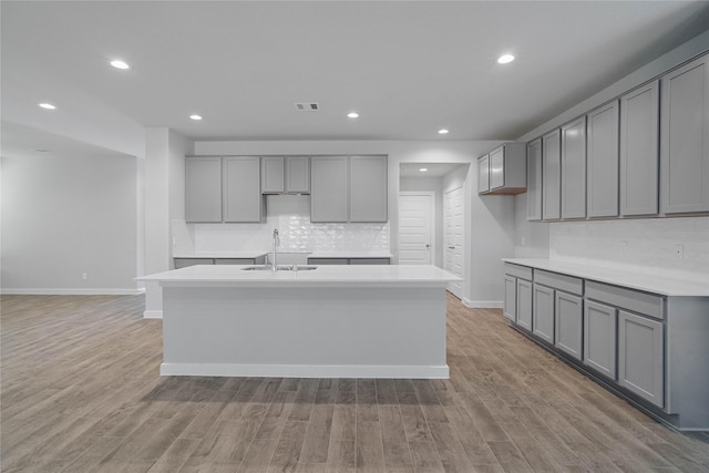 kitchen with decorative backsplash, gray cabinetry, a kitchen island with sink, sink, and light hardwood / wood-style flooring