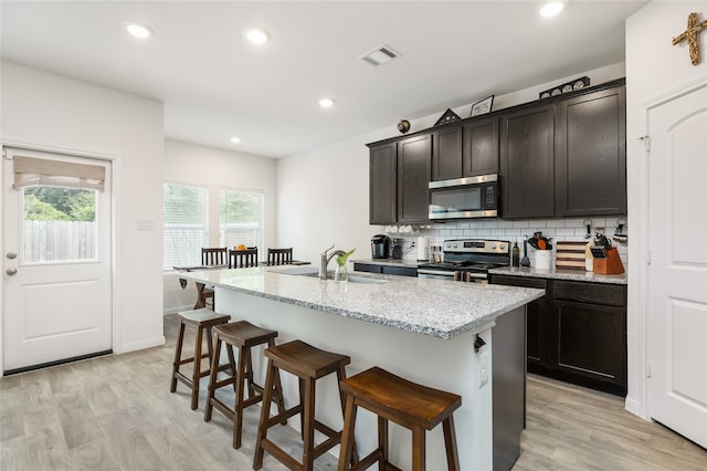 kitchen with light stone countertops, stainless steel appliances, sink, light hardwood / wood-style floors, and an island with sink