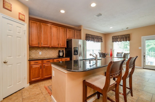 kitchen featuring sink, tasteful backsplash, dark stone countertops, stainless steel fridge, and a center island with sink