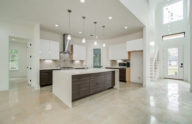 kitchen featuring wall chimney range hood, a kitchen island with sink, white cabinets, and backsplash