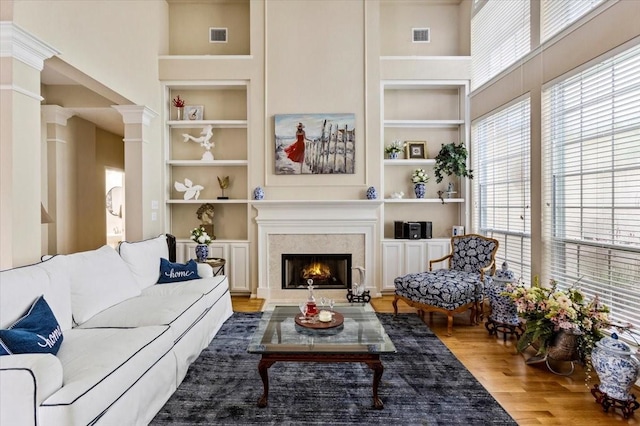 living room featuring decorative columns, built in shelves, wood finished floors, and a lit fireplace