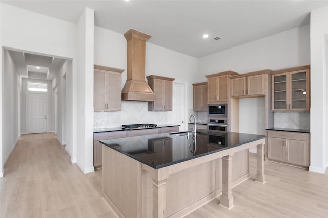 kitchen featuring sink, custom exhaust hood, a center island with sink, light wood-type flooring, and stainless steel appliances