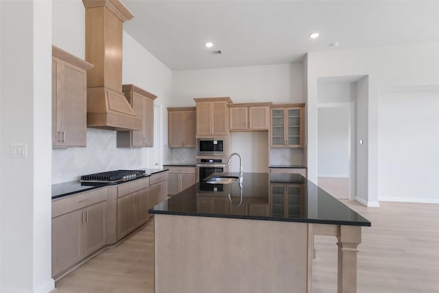 kitchen featuring appliances with stainless steel finishes, sink, custom exhaust hood, a kitchen island with sink, and light hardwood / wood-style flooring