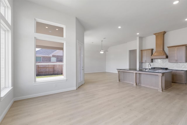 kitchen featuring sink, a center island with sink, light wood-type flooring, custom range hood, and decorative backsplash