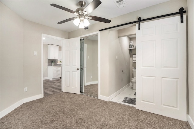 unfurnished bedroom with a barn door, ceiling fan, ensuite bathroom, and light carpet