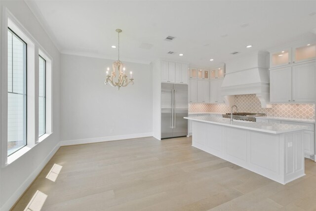 kitchen with custom range hood, white cabinets, a center island with sink, built in fridge, and backsplash