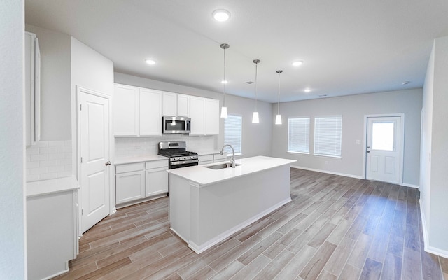 kitchen with an island with sink, hanging light fixtures, stainless steel appliances, light wood-type flooring, and white cabinetry