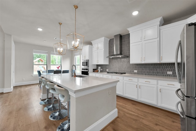 kitchen featuring wall chimney range hood, stainless steel appliances, decorative backsplash, light wood-type flooring, and a center island with sink