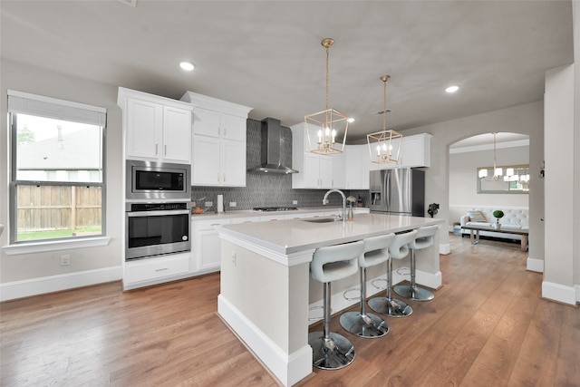 kitchen featuring wall chimney exhaust hood, sink, light hardwood / wood-style floors, appliances with stainless steel finishes, and a kitchen island with sink