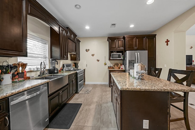 kitchen with light stone countertops, a kitchen island, dark brown cabinetry, a breakfast bar, and appliances with stainless steel finishes