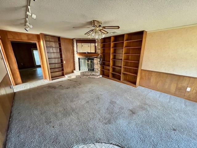 unfurnished living room with ceiling fan, rail lighting, a brick fireplace, wood walls, and a textured ceiling