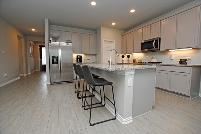 kitchen with gray cabinets, tasteful backsplash, stainless steel appliances, light stone countertops, and a center island with sink