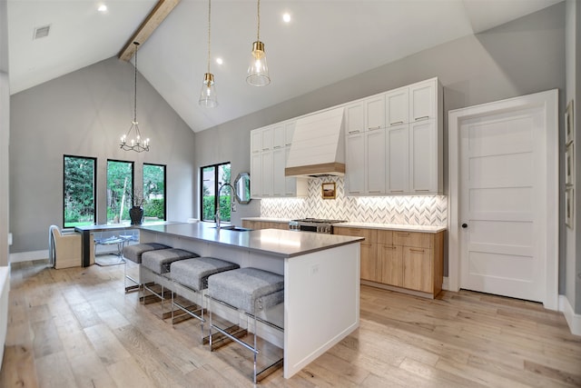 kitchen featuring white cabinetry, premium range hood, high vaulted ceiling, a center island with sink, and light hardwood / wood-style flooring