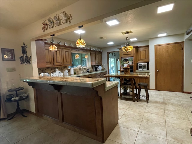 kitchen featuring kitchen peninsula, hanging light fixtures, stainless steel microwave, tile counters, and light tile patterned flooring