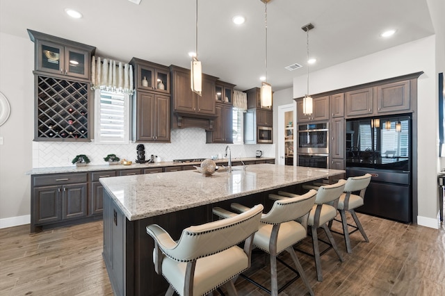 kitchen with decorative light fixtures, wood-type flooring, stainless steel appliances, decorative backsplash, and a center island with sink