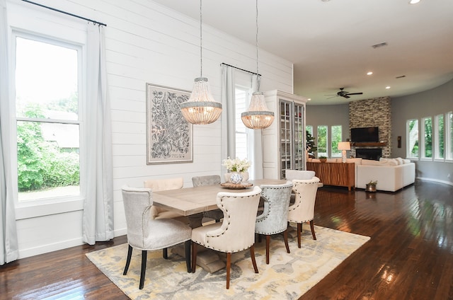 dining room with a stone fireplace, dark wood-type flooring, and ceiling fan with notable chandelier