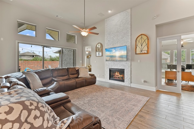 living room with ceiling fan, a stone fireplace, light wood-type flooring, and a high ceiling