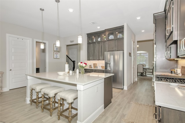 kitchen with stainless steel fridge with ice dispenser, tasteful backsplash, light wood-type flooring, a kitchen island with sink, and hanging light fixtures