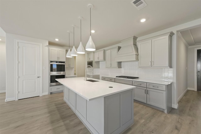 kitchen with sink, a kitchen island with sink, double oven, tasteful backsplash, and custom range hood