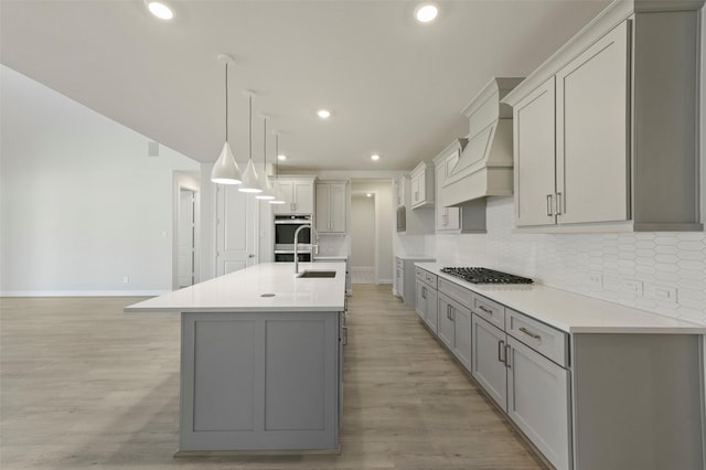 kitchen featuring premium range hood, decorative light fixtures, tasteful backsplash, gray cabinetry, and a kitchen island with sink