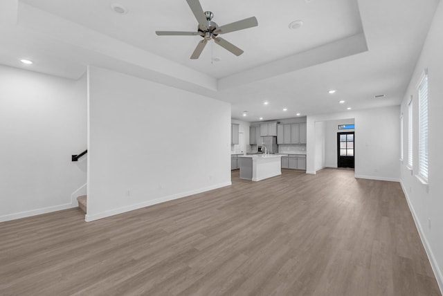 unfurnished living room with light wood-type flooring, sink, ceiling fan, and a raised ceiling