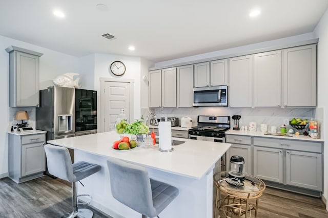 kitchen with a breakfast bar area, dark hardwood / wood-style floors, appliances with stainless steel finishes, backsplash, and a kitchen island with sink